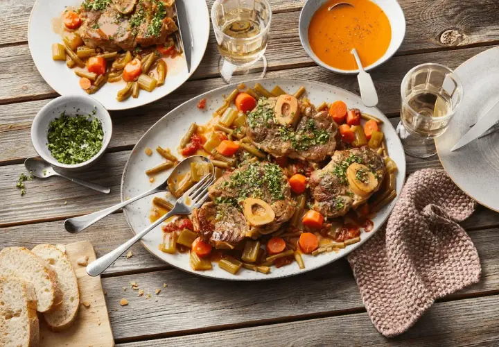 Ein festliches Mahl: saftiges Ossobuco mit Gemüse und einem Glas Weißwein. Dazu frisches Brot und eine cremige Tomatensuppe.
