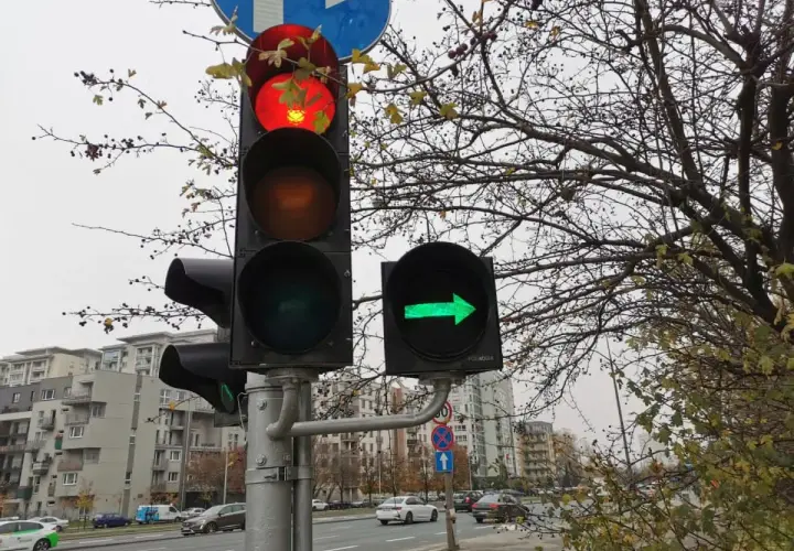 Sygnalizator S2 z czerwonym światłem na górze i zieloną strzałką w prawo. Za nim drzewa i budynki.