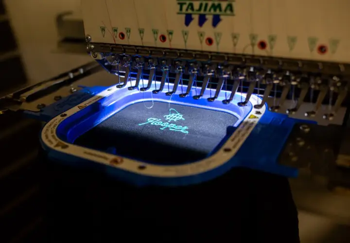 Machine à broder Tajima créant une estampille digitalisée sur un tissu noir. Le logo "Hooper" est en cours de réalisation avec du fil turquoise.
