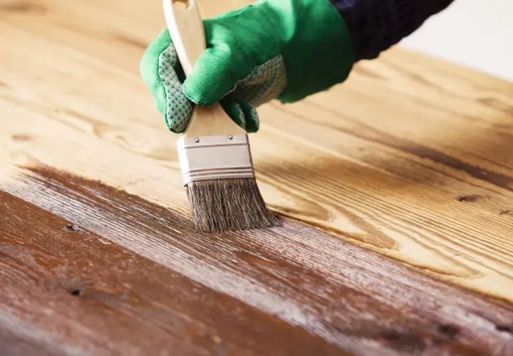 Mano con guante verde aplicando barniz sobre madera, creando un acabado brillante. Barnizar sobre barniz sin lijar para renovar la superficie.