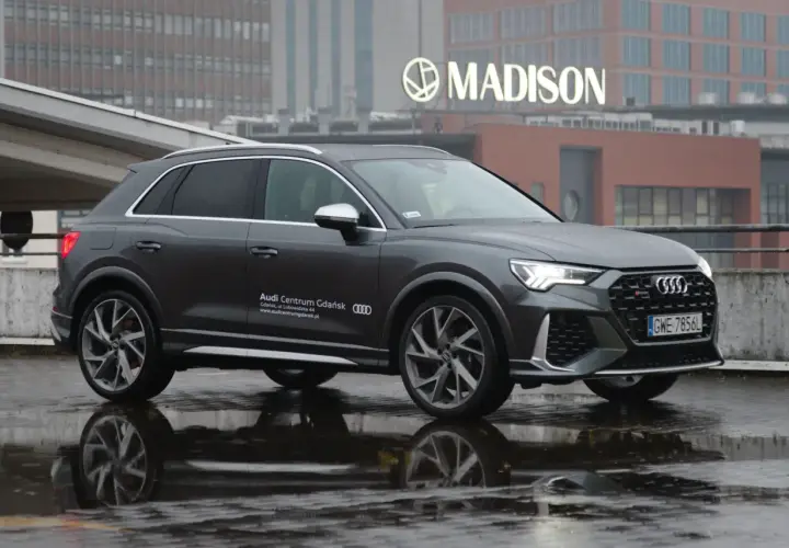 Szary Audi RS Q3 na mokrej nawierzchni, z odbiciem w kałuży. W tle budynek z napisem MADISON.
