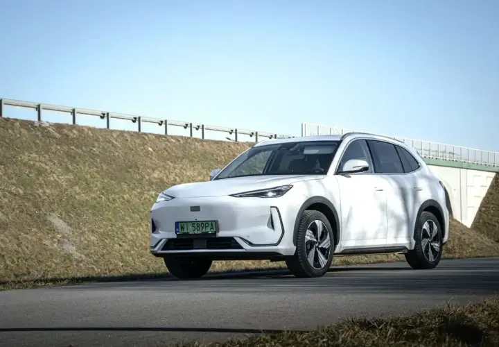 Biały Geely EX5 na tle słonecznego nieba i zielonych wzgórz. Elegancki SUV gotowy na podróż.