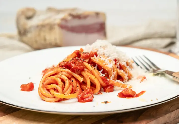 Ein Teller Pasta all'Amatriciana, garniert mit geriebenem Parmesan. Im Hintergrund ein Stück Guanciale, die Zutat für das authentische Amatriciana Rezept.
