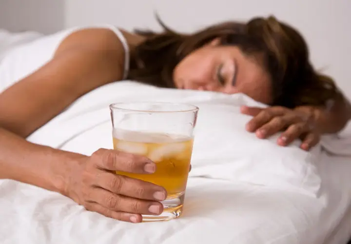 Kobieta śpi w łóżku, trzymając szklankę z napojem i lodem. Może to być alkohol, np. GHB.