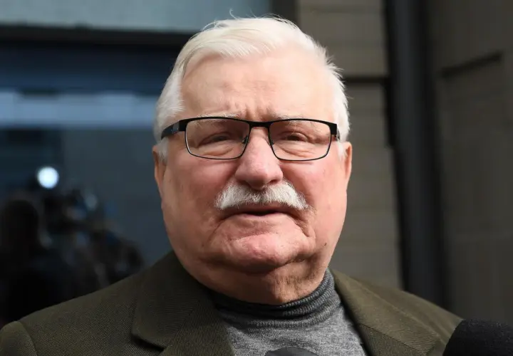 Lech Wałęsa w okularach i z wąsami, w swetrze i marynarce. W tle niewyraźne postacie.