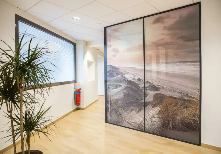 Un couloir moderne avec une plante verte et un extincteur, séparé par une grande cloison amovible ornée d'un paysage de dunes et de plage.