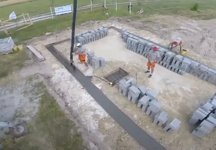 Pracownicy wylewają beton na fundamenty. Widać koparkę, betoniarkę i sterty pustaków.
