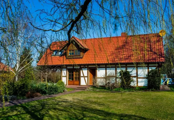 Urokliwy dom z czerwonym dachem, otoczony zielenią. Idealne miejsce na luksusową agroturystykę, gdzie można odpocząć od miejskiego zgiełku.