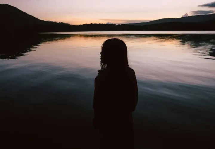 Sylwetka dziewczynki nad jeziorem o zachodzie słońca. Woda odbija niebo, tworząc spokojny obraz. Idealne miejsce na chwilę refleksji, jak w cytaty o spokoju i ciszy.