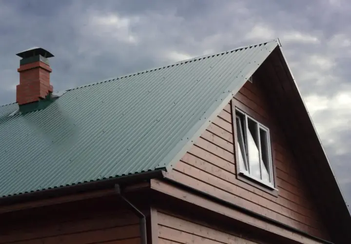 Dach domu pokryty zieloną blachą trapezową, widoczny komin i okno. Szerokość krycia blachy trapezowej zapewnia szczelność.
