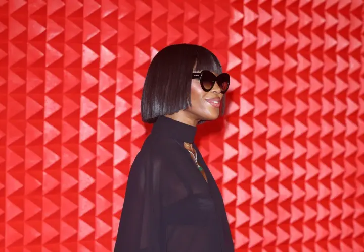 Naomi Campbell luce un corte de pelo **blunt cut** y gafas de sol, posando ante un fondo rojo texturizado.