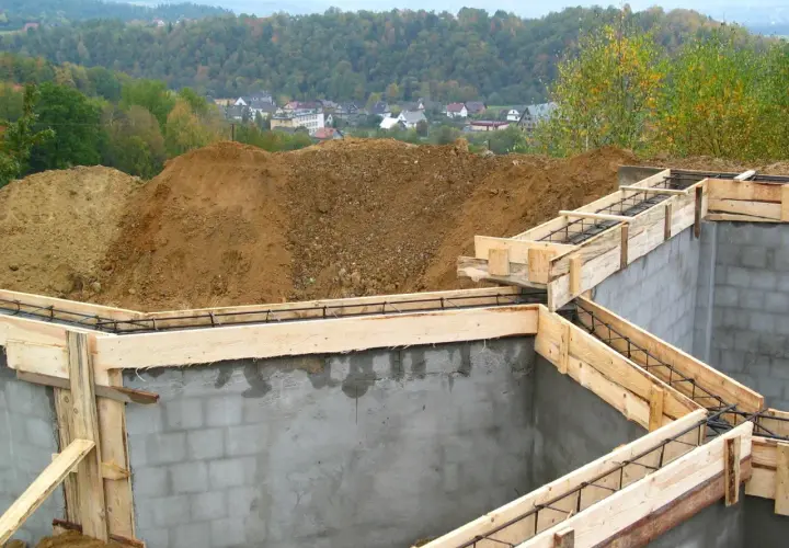 Fundament budowy z szalunkiem i zbrojeniem, w tle domy i las.