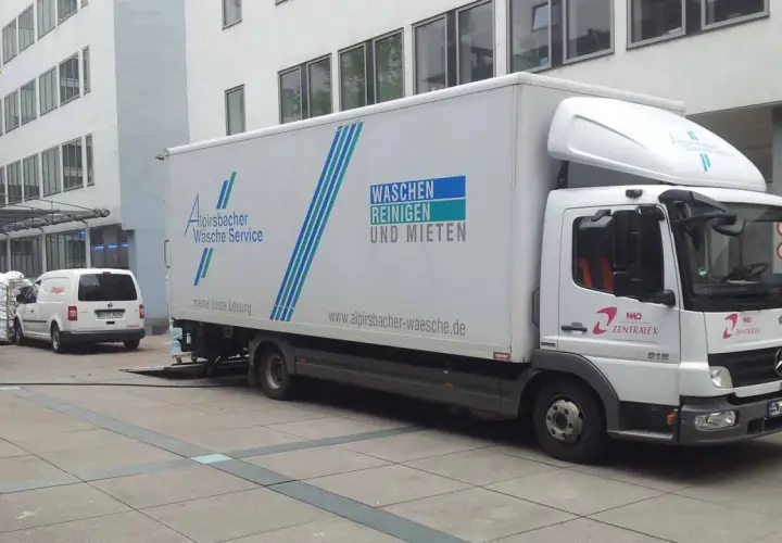Ein LKW von Alpirsbacher Wäsche Service, der für Werkverkehr genutzt wird, steht vor einem Gebäude. Er transportiert Wäsche.