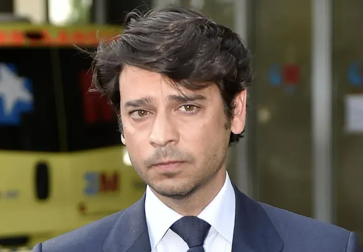 Miguel Palomo Danko, con traje oscuro y corbata, mira fijamente. Detrás, una ambulancia amarilla.