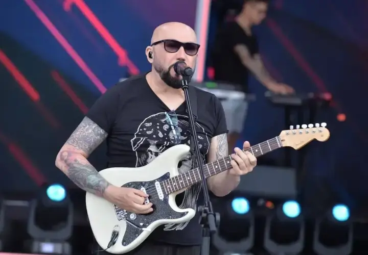 Artysta z tatuażami gra na białej gitarze, śpiewając do mikrofonu. W tle widać klawiszowca. To z pewnością niezapomniane **kombi utwory** na żywo!