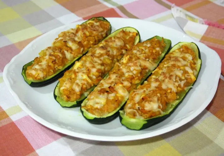 Cuatro calabacines rellenos de atún, gratinados con queso, servidos en un plato blanco sobre un mantel a cuadros.