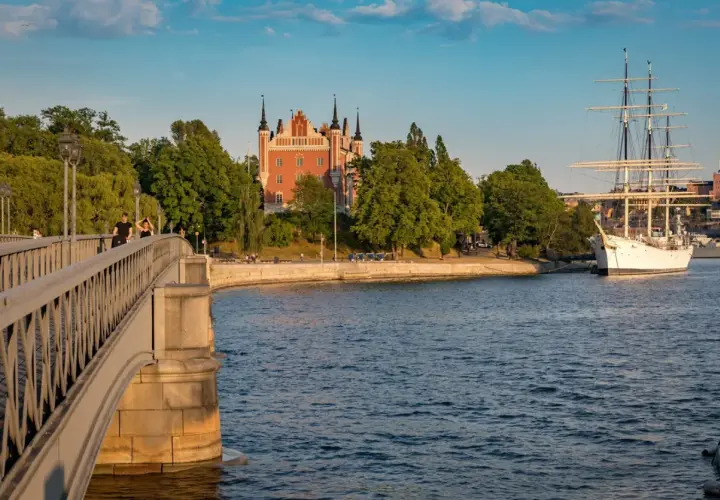 En vacker scen i Stockholm med en bro i förgrunden, ett rött tornliknande hus och ett stort vitt segelfartyg i vattnet.