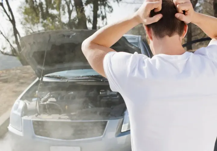 Hombre frustrado mira cómo sale humo blanco del motor de su coche averiado.