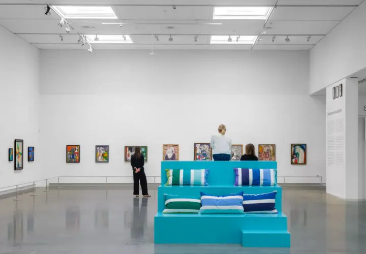 I en konsthall sitter besökare på en turkos scen med kuddar, omgivna av färgglada tavlor. Kanske är de där för att inspireras, för att vara en musa för konstnären.