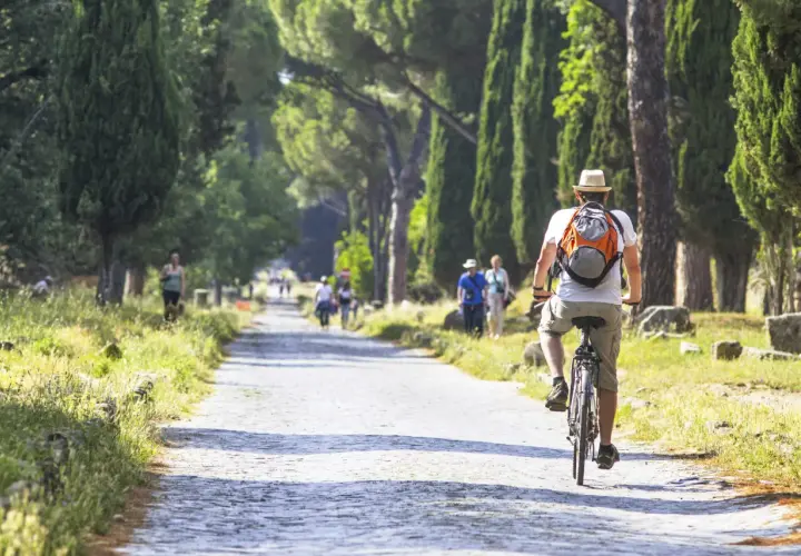 Un hombre pedalea por un camino empedrado en Roma, rodeado de altos cipreses y ruinas antiguas.
