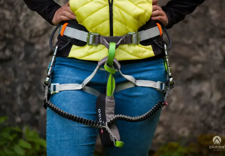 Osoba w kamizelce i jeansach ma na sobie sprzęt do via ferrata, w tym lonżę z karabinkami.