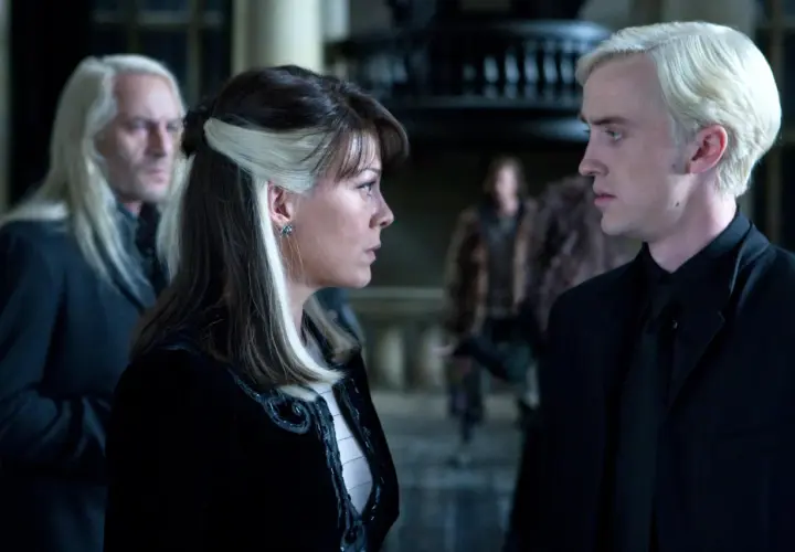 Narcyza Malfoy rozmawia z synem, Draco, w ponurym wnętrzu. W tle widać Lucjusza Malfoya.