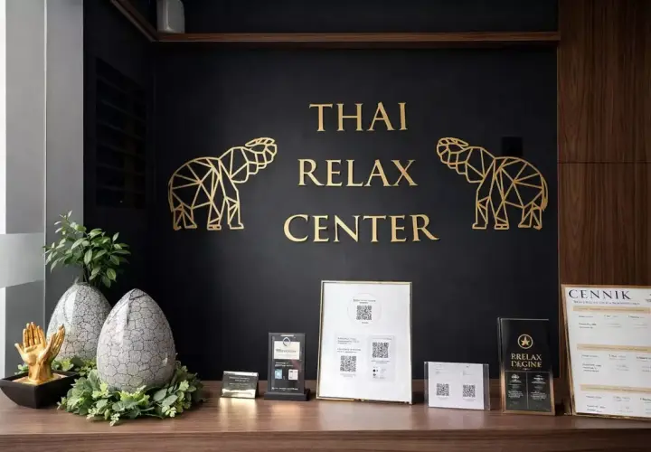 Recepcja centrum masażu tajskiego. Złote słonie i napis "THAI RELAX CENTER" na czarnej ścianie. Na blacie dekoracje i cennik. Tu możesz się wyciszyć.