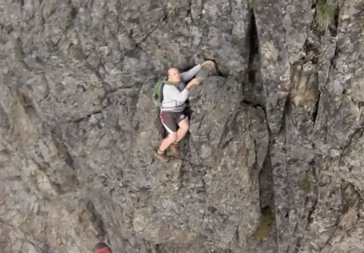 A man is climbing a steep, rocky cliff face. He's wearing shorts and a backpack, looking like he's truly "cliffed out" and facing a challenging ascent.