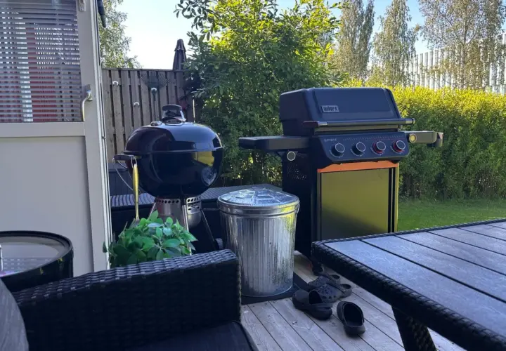 En Weber kolgrill bäst i test står redo för sommarens grillkvällar bredvid en gasolgrill. Trädgårdsmöbler och en fin gräsmatta skapar en härlig atmosfär.