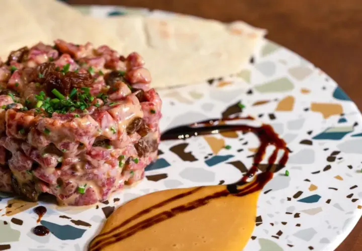 Delicioso tartar de salchichón, con trocitos de frutos secos y cebollino fresco, servido en un plato moderno con salsas.