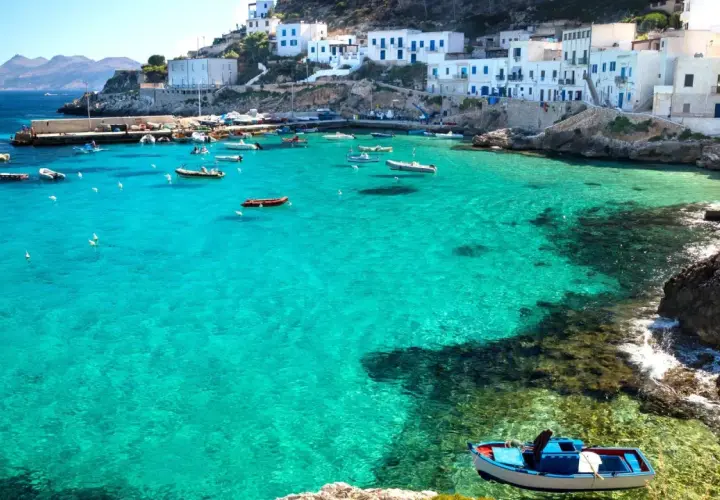 Boats in a turquoise bay near white buildings, a picturesque scene reminiscent of the charm found near international airports in Sicily.
