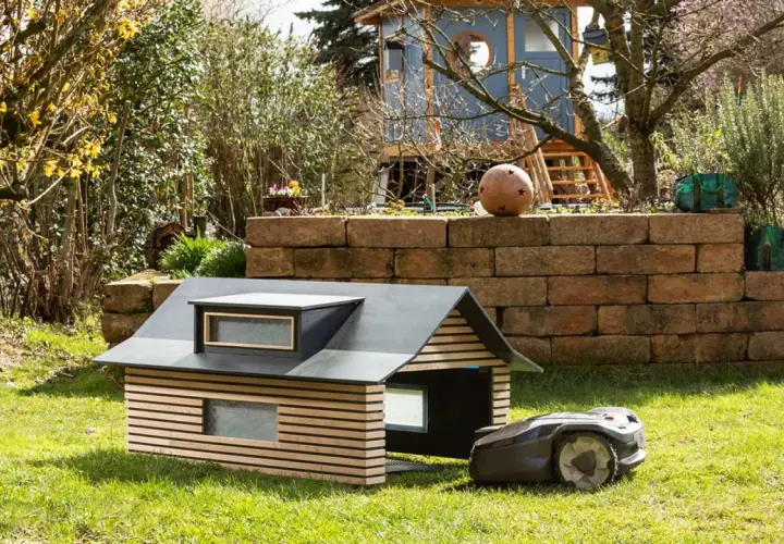 Ein moderner, selbstgebauter Rasenroboter-Unterstand mit Holzlamellen und Dachfenstern. Der Rasenroboter parkt daneben.
