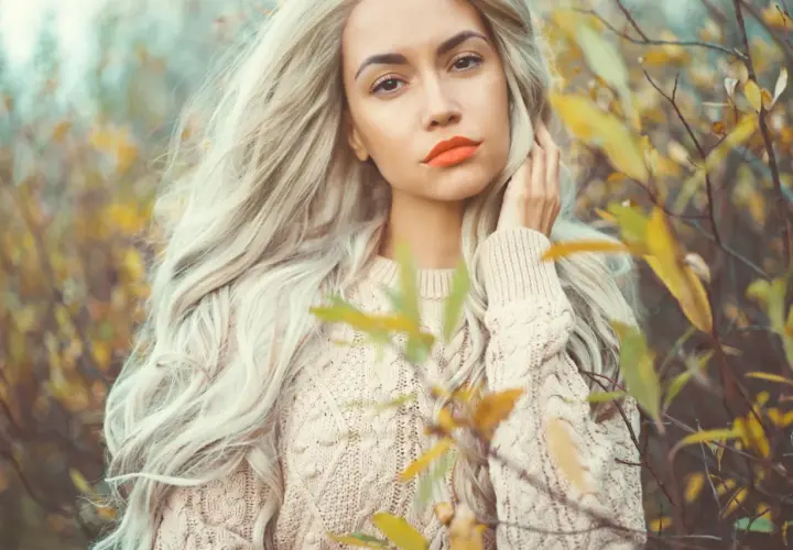 Kobieta z burzą waniliowy blond włosów i pomarańczowymi ustami w beżowym swetrze, otoczona jesiennymi liśćmi.
