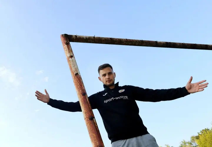 Młody piłkarz z Częstochowy, w czarnej bluzie z logo "sport-active", pozuje na tle nieba i zardzewiałej poprzeczki bramki.
