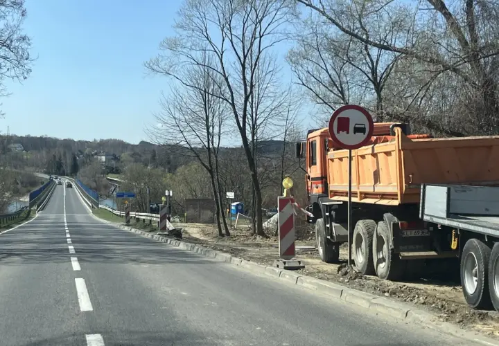 Pomarańczowa wywrotka MAN stoi przy drodze, gdzie trwa remont. Znak zakazu wyprzedzania dla pojazdów ciężarowych. Velo Raba w tle.