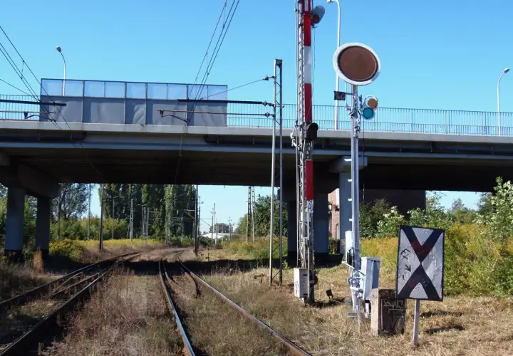 Tarcza ostrzegawcza przy torach kolejowych pod wiaduktem. Sygnalizacja świetlna i znak krzyżowy informują o zbliżającym się niebezpieczeństwie.