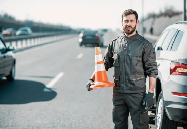 Pomoc drogowa na autostradzie. Mężczyzna w roboczym ubraniu trzyma pomarańczowy pachołek, gotowy do zabezpieczenia miejsca awarii auta.