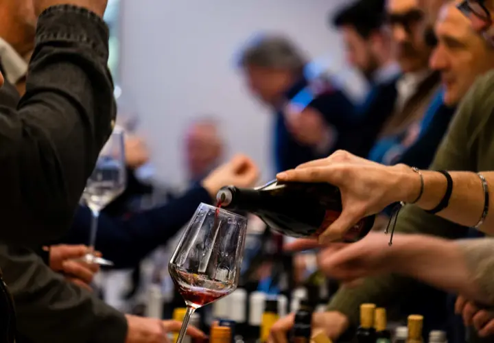 Versando vino rosso in un calice, si apprezzano le caratteristiche del vermentino. Persone in attesa, bottiglie sullo sfondo.