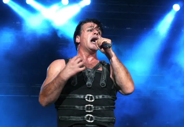 Till Lindemann na koncercie, w czarnym, skórzanym stroju, śpiewa do mikrofonu na tle niebieskich świateł.