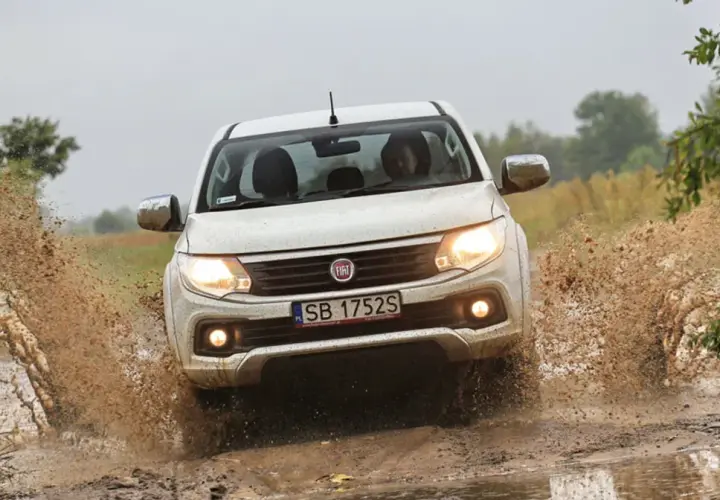 Biały Fiat Fullback z impetem wjeżdża w błotnistą kałużę, wzbijając fontanny brudu.