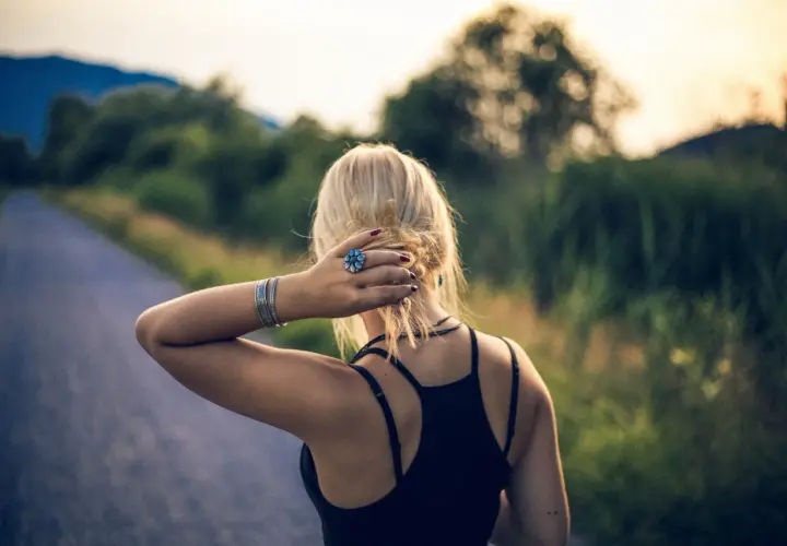 Blondynka z pierścionkiem w kształcie kwiatka, jak mały szelma, poprawia włosy na tle drogi i zieleni.
