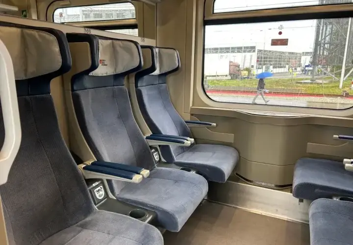 Wygodny wagon z przedziałami, z niebieskimi siedzeniami i stolikami. Za oknem deszczowa pogoda i osoba z parasolem.