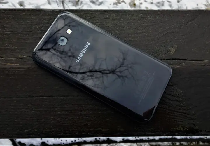 Czarny Samsung SM-A520F leży na drewnianej ławce, odbijając gałęzie drzew.