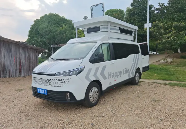 Ce camping-car 9m blanc, avec "Happy Life" écrit dessus, est prêt pour l'aventure. Il est garé sur un chemin de gravier, à côté d'une structure en bois.