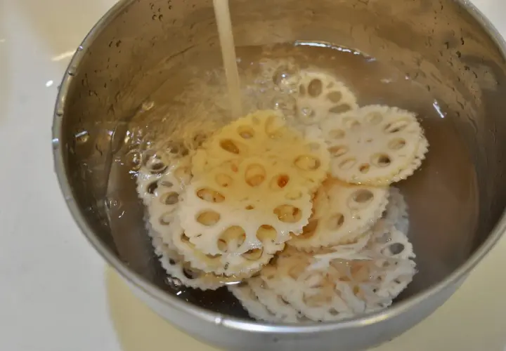 Slices of pickled lotus root are being rinsed in a metal bowl under running water, ready for a delicious meal.