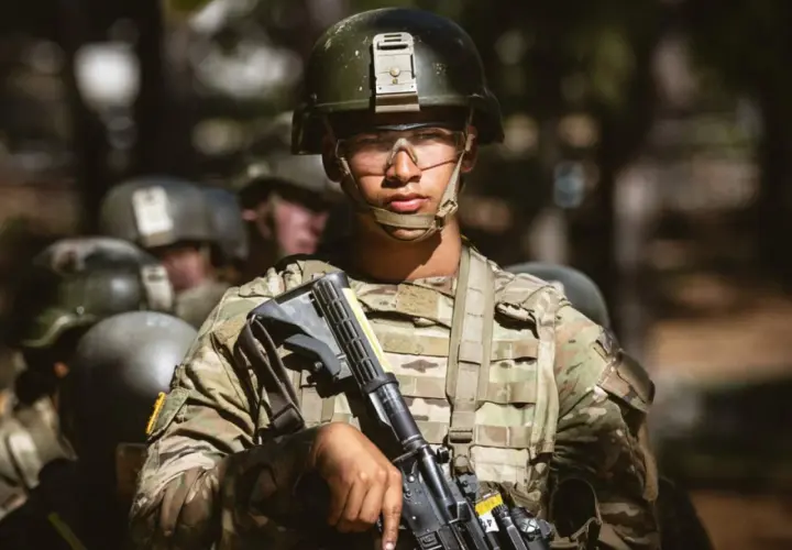A young soldier, determined and focused, stands ready during far bootcamp. He wears a helmet, protective eyewear, and camouflage fatigues, holding his rifle.