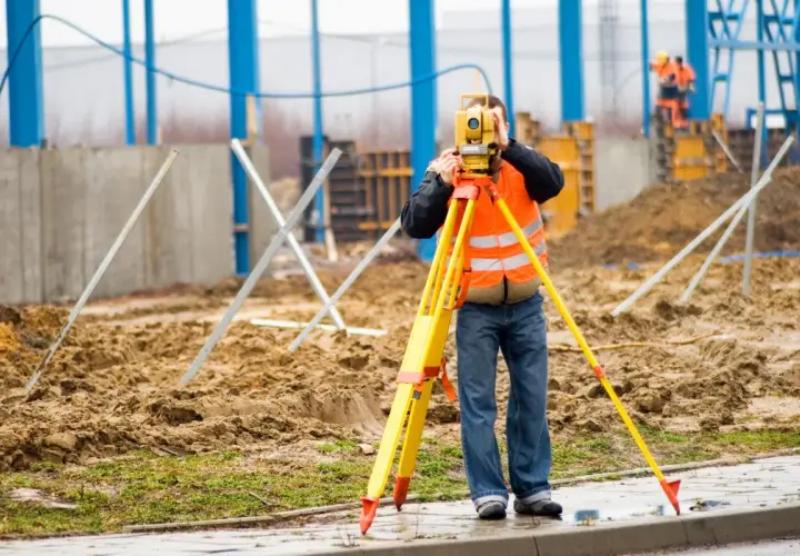 Geodeta w kamizelce odblaskowej mierzy teren za pomocą niwelatora na budowie.