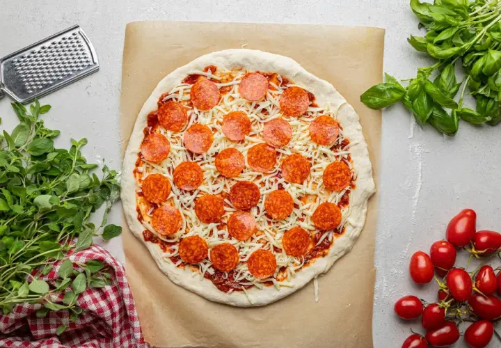 Pizza z sosem, serem i okrągłymi plasterkami pepperoni, gotowa do pieczenia. Obok świeża bazylia i pomidorki.