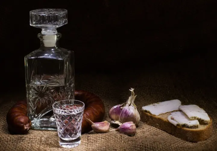Wódka smakowa w karafce i kieliszku, obok kiełbasa, czosnek i chleb ze smalcem. Widoczne objawy utlenienia wódki smakowej po długim staniu w słońcu.