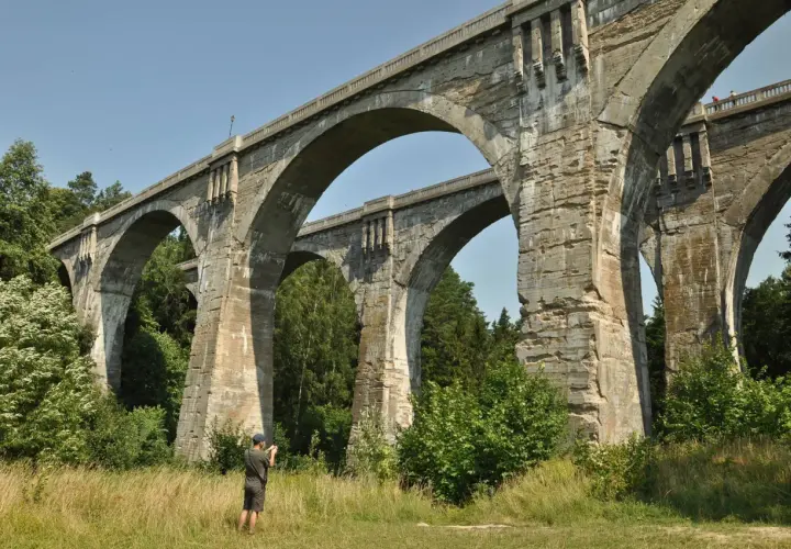 Imponujące mosty w Stańczykach, zbudowane z kamienia, górują nad zielonym krajobrazem. Mężczyzna z aparatem dokumentuje ich monumentalność.